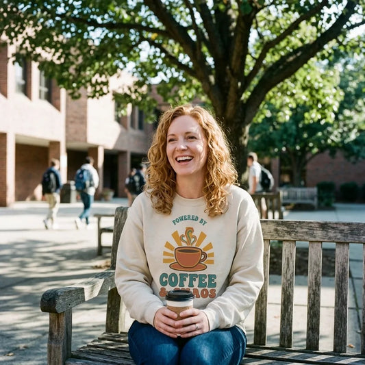 Smiling teacher in a Sand Powered By Coffee And Chaos crewneck sitting on a school courtyard bench holding coffee