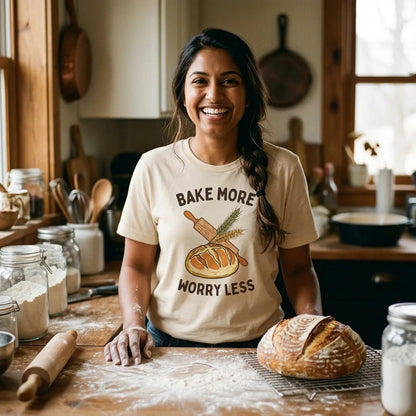 Smiling home baker in a farmhouse kitchen with a fresh sourdough loaf, wearing the Bake More Worry Less sourdough tee