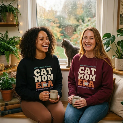 Two friends laughing in Cat Mom Era crewnecks (Black and Maroon) with a grey cat by a rainy window — funny cat mom sweatshirt