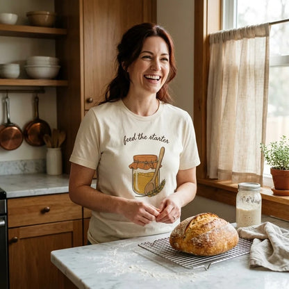 Feed The Starter sourdough baker tee in Natural on a smiling woman in a cozy home kitchen with a fresh sourdough loaf