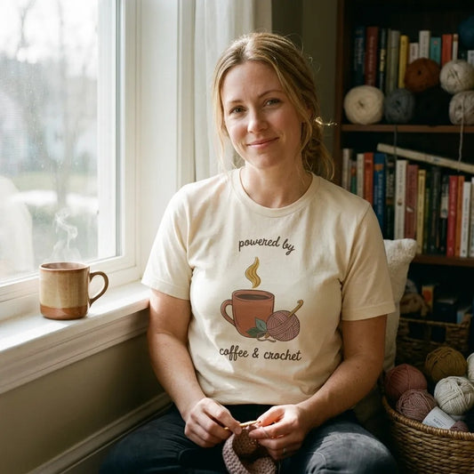 Powered by Coffee and Crochet cottagecore crochet tee — worn by a woman relaxing in a sunny window reading nook with yarn and coffee (Natural)