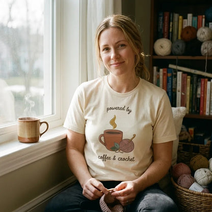 Powered by Coffee and Crochet cottagecore crochet tee — worn by a woman relaxing in a sunny window reading nook with yarn and coffee (Natural)