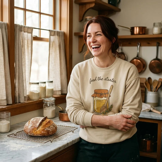 Feed The Starter sourdough crewneck — woman laughing in a cozy home kitchen, Sand sweatshirt